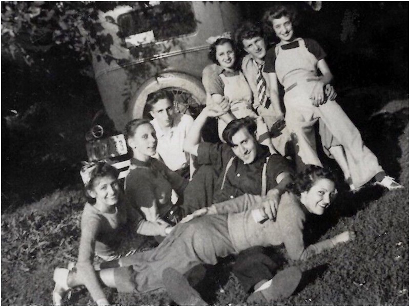 a group of college students sitting on the grass behind a vintage touring car while one of their number lies over her boyfriend's lap for a spanking